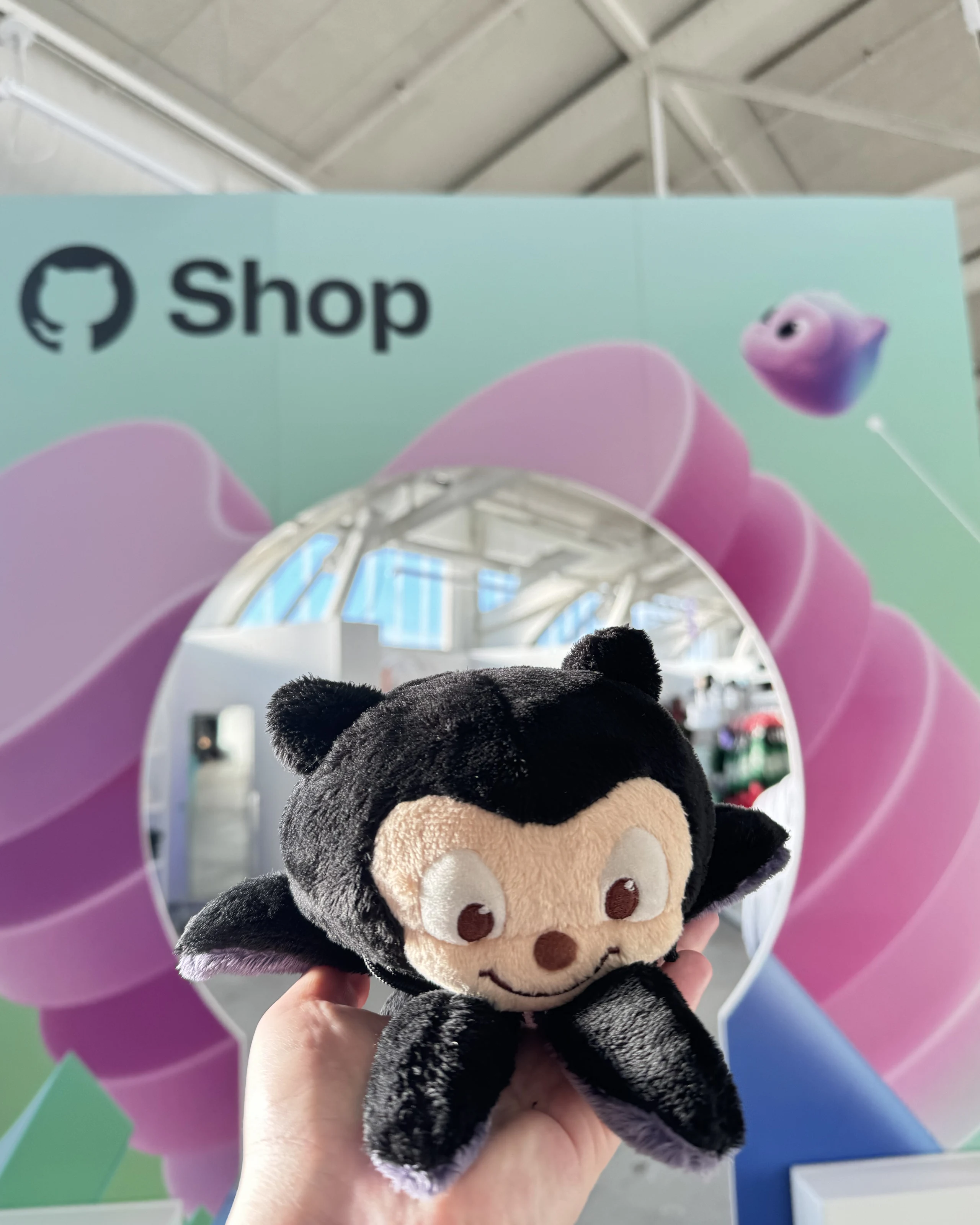 Mona the octocat plushie held up by a hand in front of the GitHub Shop entrance at GitHub Universe 2024.