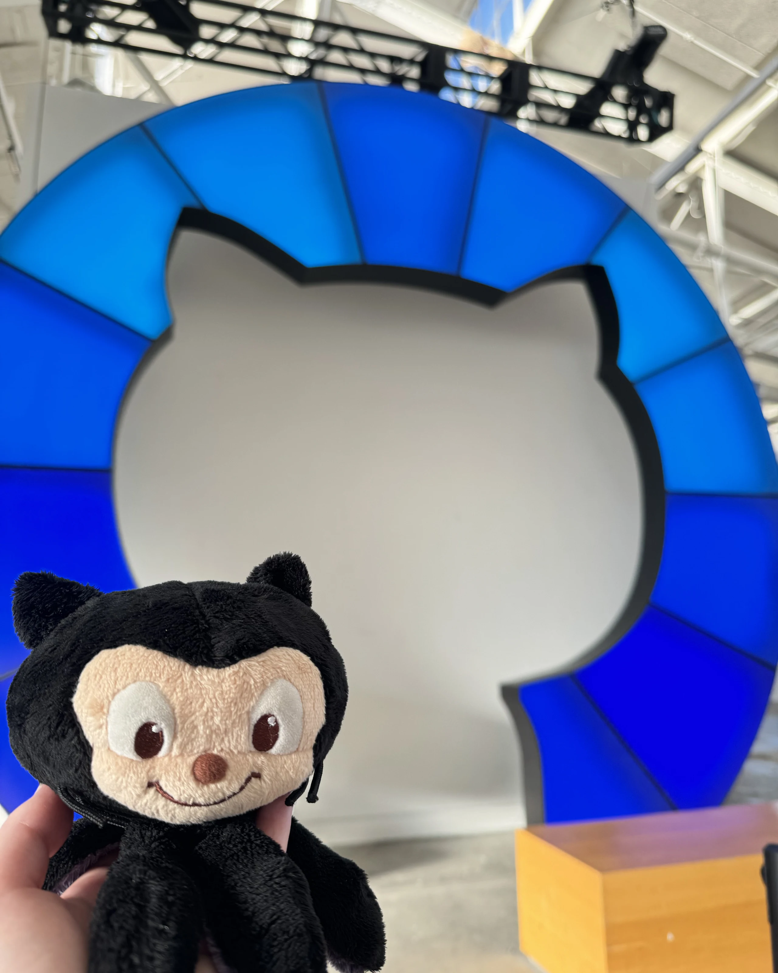 Mona the octocat plushie held up by a hand in front of a blue GitHub logo installation.